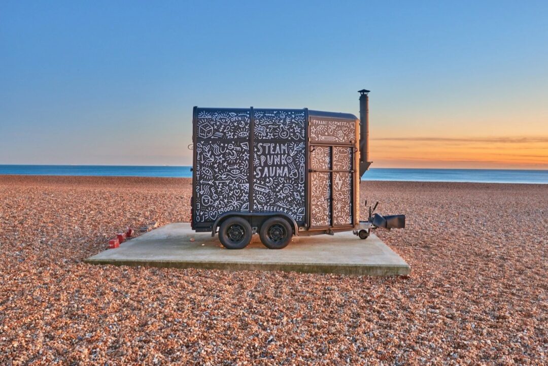 Portable sauna on a seaside pebble beach at sunset, with “STEAM” lettering on the side.