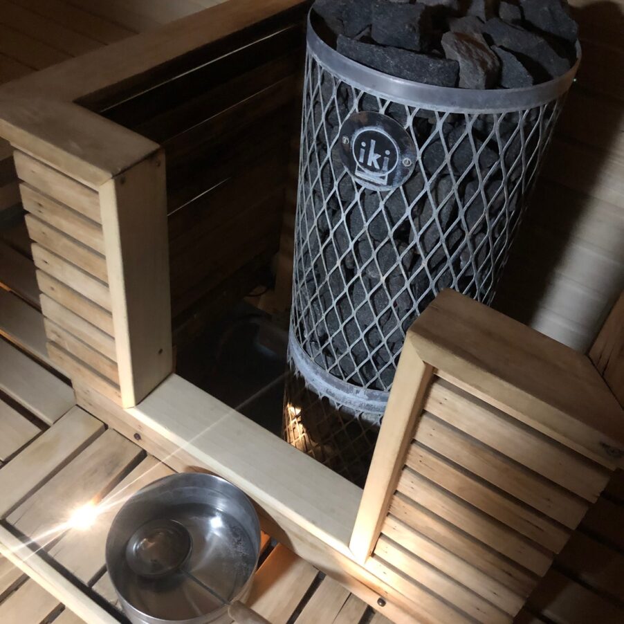 Sauna stove with hot stones, water bucket and ladle in a wooden UK sauna; “iki” logo on the heater.