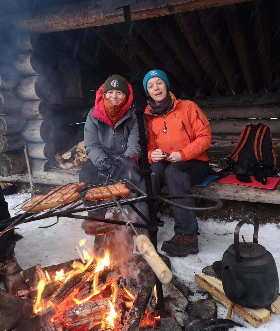 Two women warm by a campfire, roasting food beside a snowy log cabin in winter