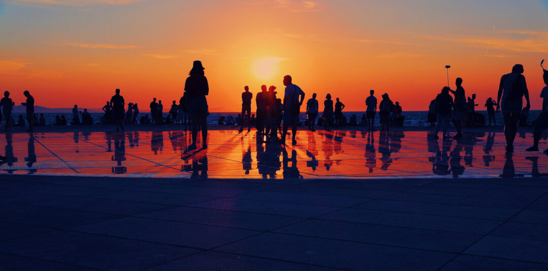 People walking along Zadar waterfront at sunset, with silhouettes reflected in the pavement