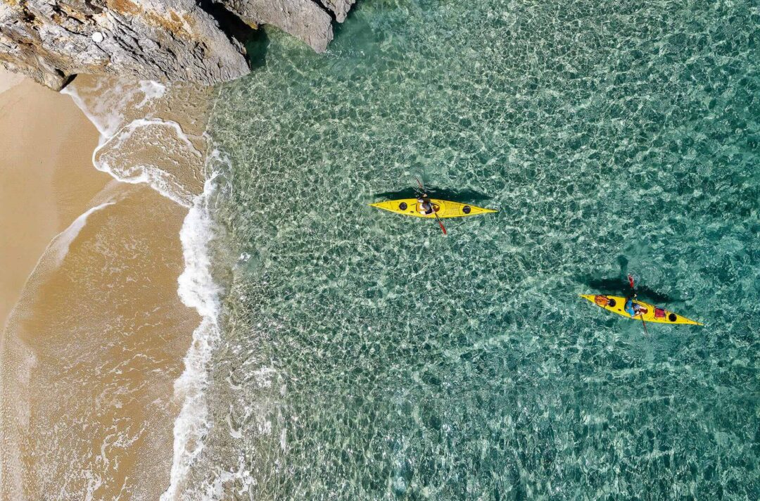 Two yellow kayaks paddling in clear coastal water near a sandy shoreline