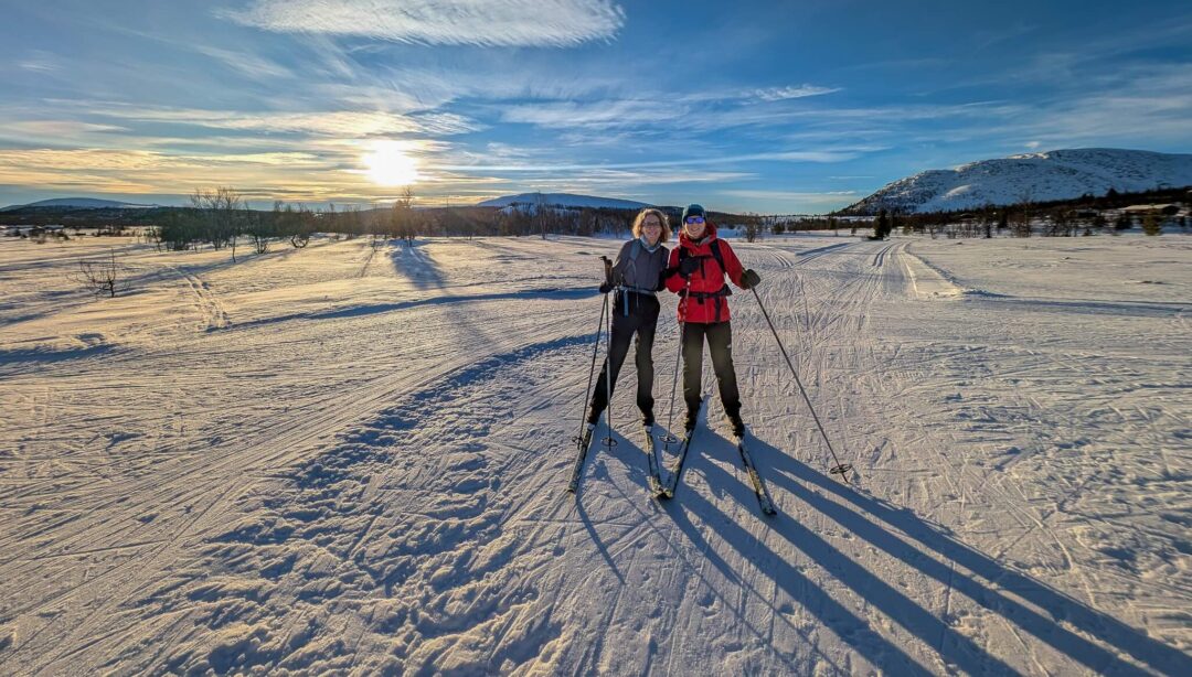 Two women cross-country skiing on a snowy track in a winter landscape at sunrise