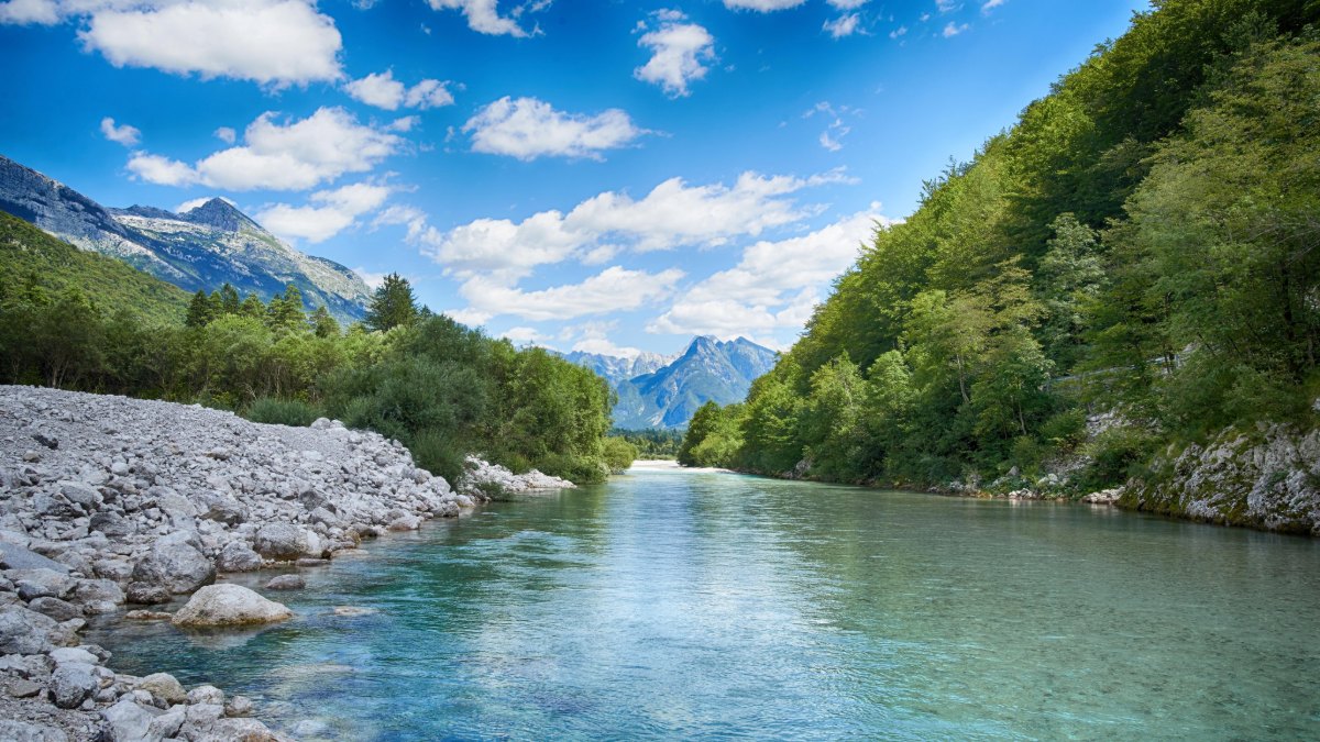 Calm river with clear blue water, rocky banks and forested mountains under a blue sky