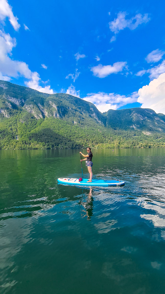 Woman standing on a stand-up paddleboard on a lake in Slovenia, with mountains and blue sky.