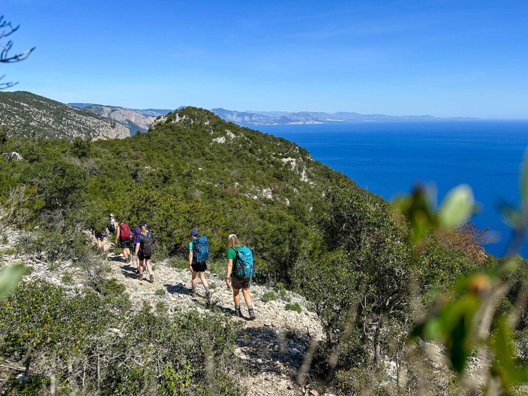 Women hike along a coastal trail with sea views across a Mediterranean hillside landscape