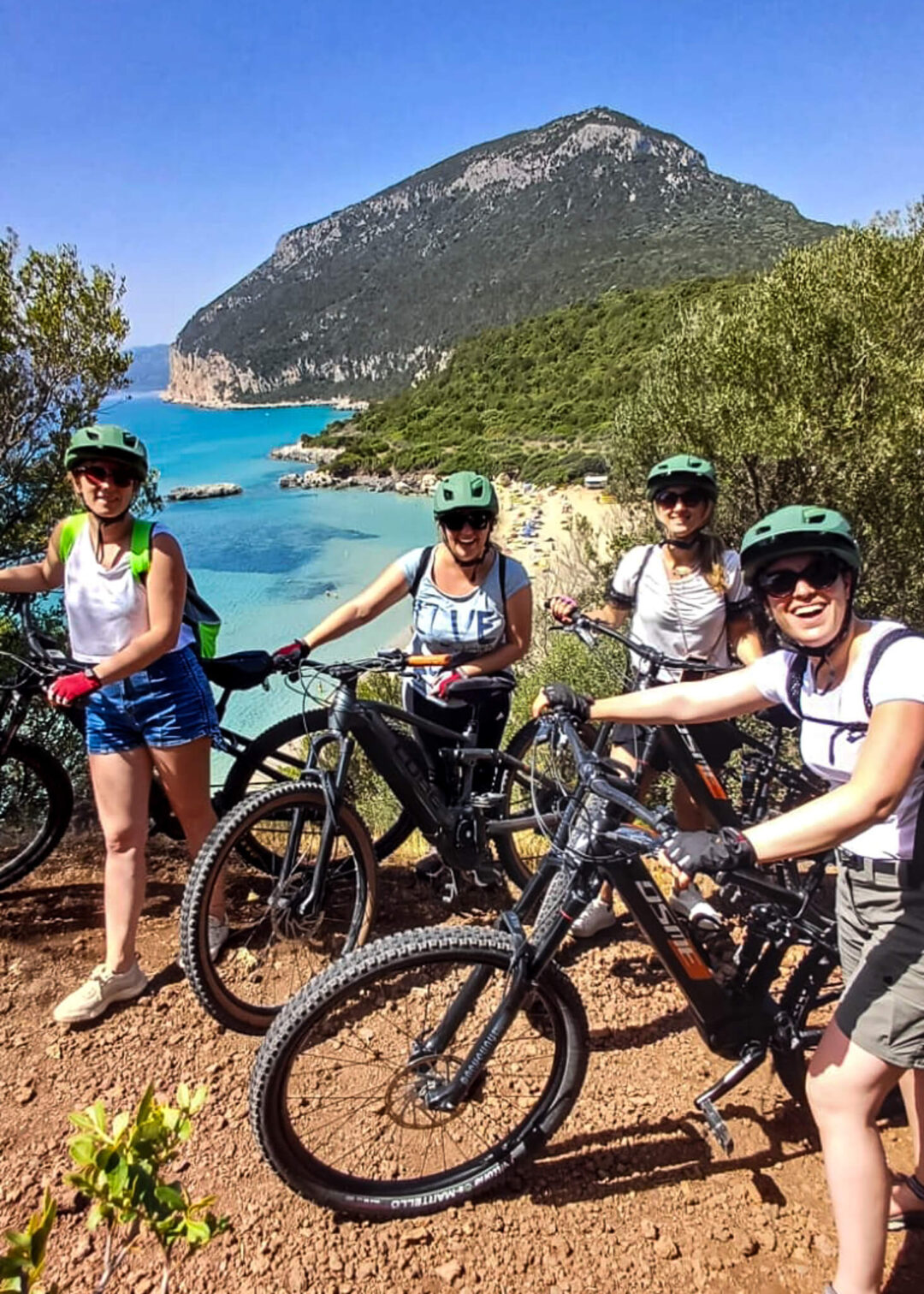 Women on eBikes on a coastal trail overlooking turquoise sea in East Sardinia