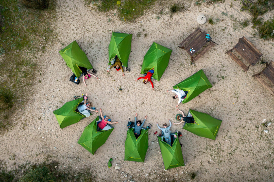 Women gathered around green tents on a sandy coastal campsite, outdoors adventure hike setting