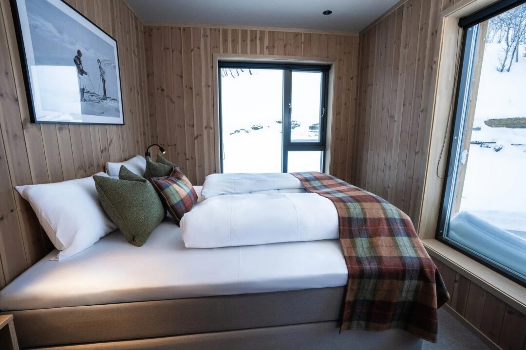 Ski lodge bedroom with double bed and plaid throws, looking out on snowy Norway through large windows