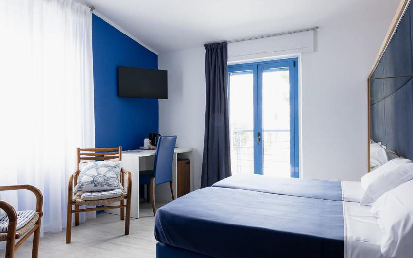 Bright hotel bedroom with a double bed, blue window frame, and TV on the wall