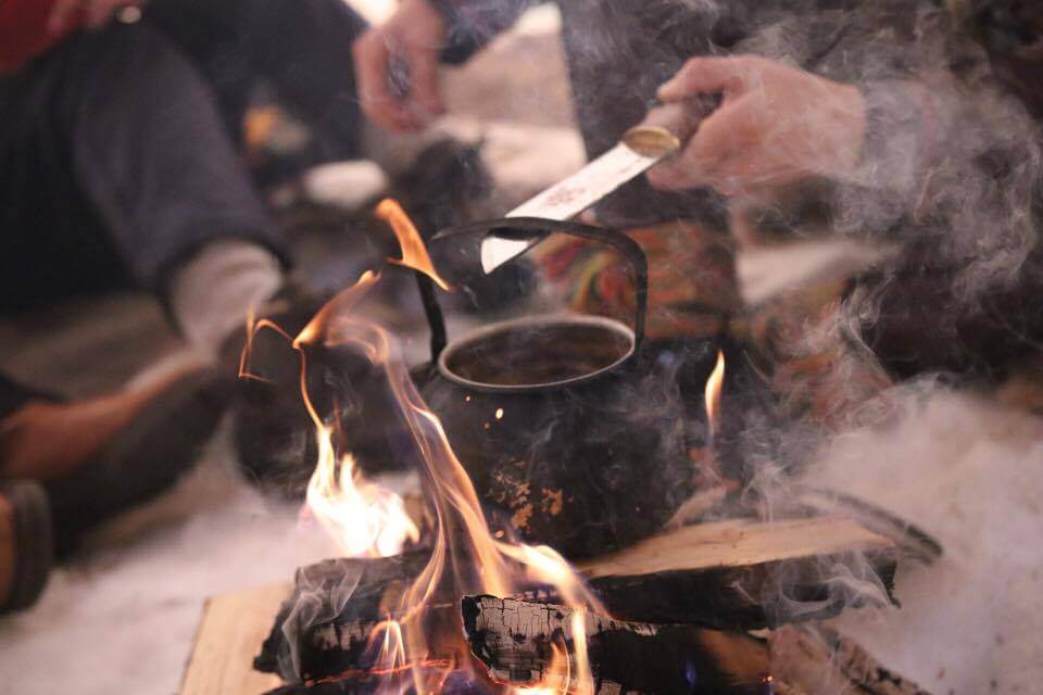Outdoor campfire cooking over flames with smoke, with hands feeding food into a fire in winter wilderness