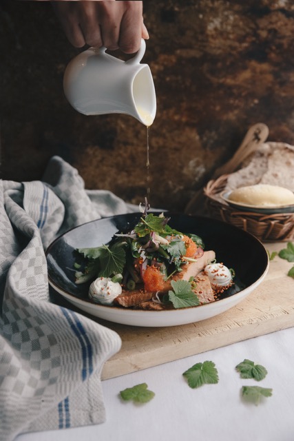 Woman pours sauce over a plated meal on a table with herbs and bread, in a winter setting