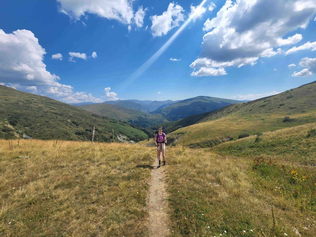 Woman hiking a mountain trail through rolling green hills under a blue sky with clouds