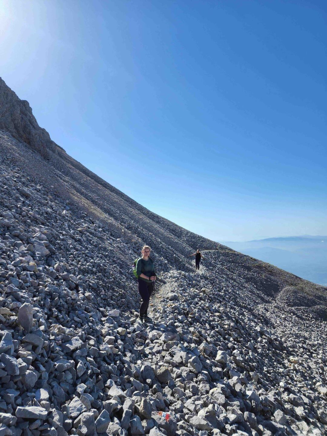 Woman hiking a rocky mountain trail under a clear blue sky, with the sea in the distance.