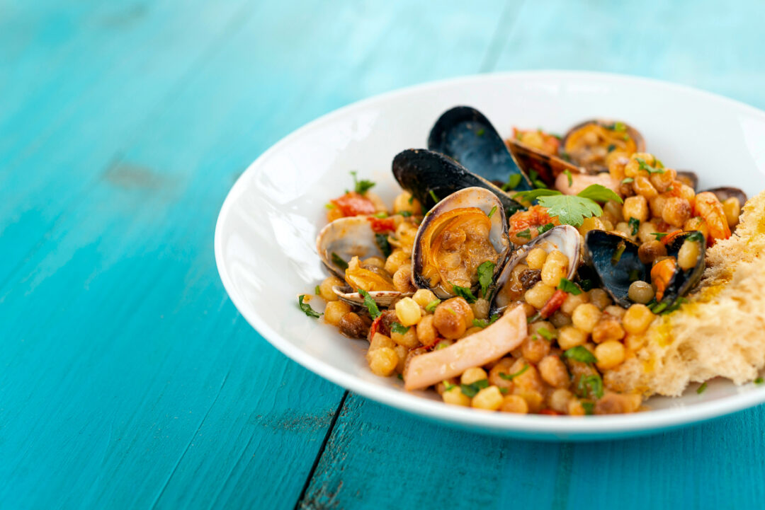 Bowl of mussels and chickpea seafood stew with bread on a blue table—Sardinia-style food.