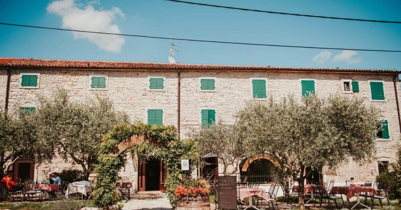 Stone courtyard with olive trees, outdoor seating and green-shutter windows, Mediterranean-style building exterior
