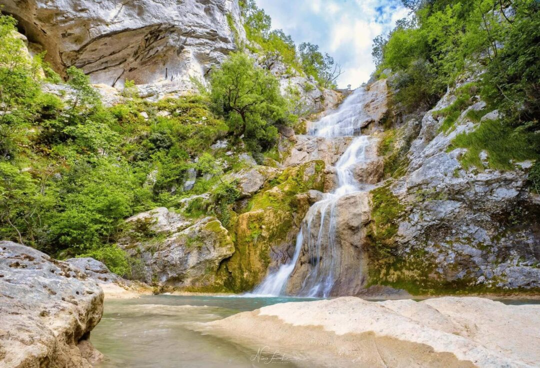 Waterfall cascading through a rocky gorge with lush green trees and stream in the background