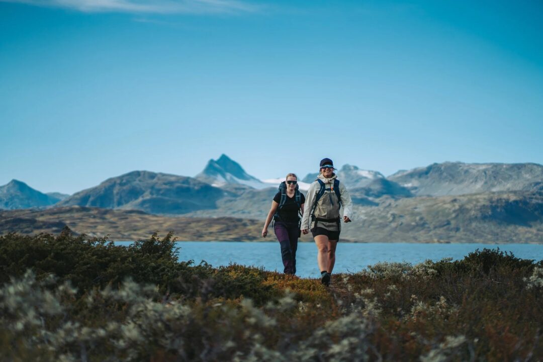 Women hiking with backpacks on a trail, with mountain and lake scenery in the background