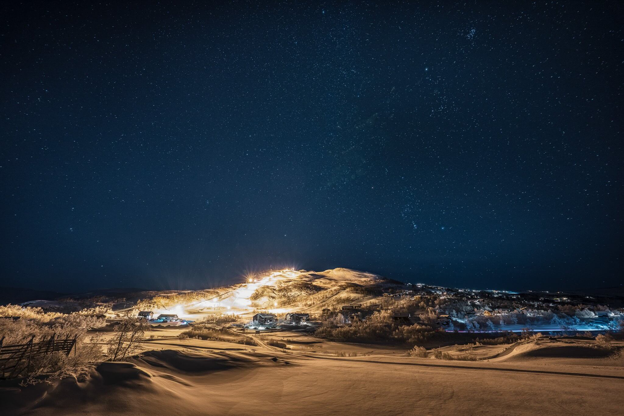 Snowy ski slopes at night under a starry sky, with resort lights and tracks