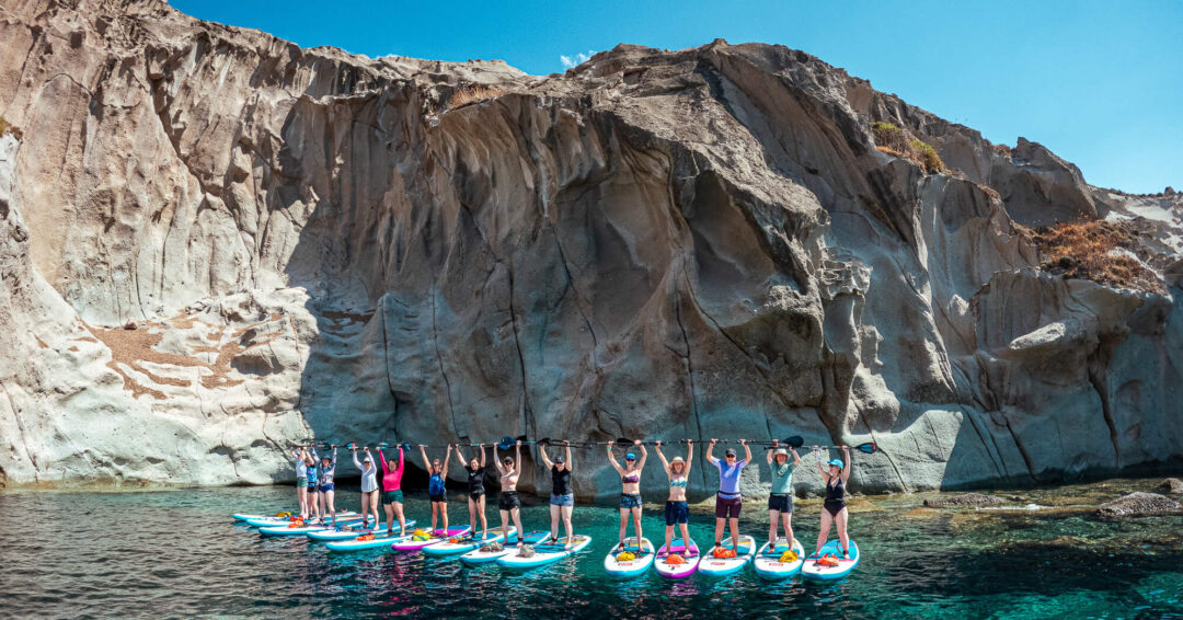 Women on stand-up paddleboards in a clear sea off a rugged cliff coast