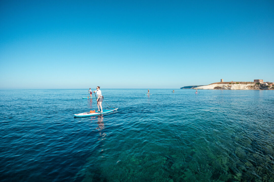 Woman paddleboarding on calm sea off the Sardinia coastline, with coastal buildings in the distance.