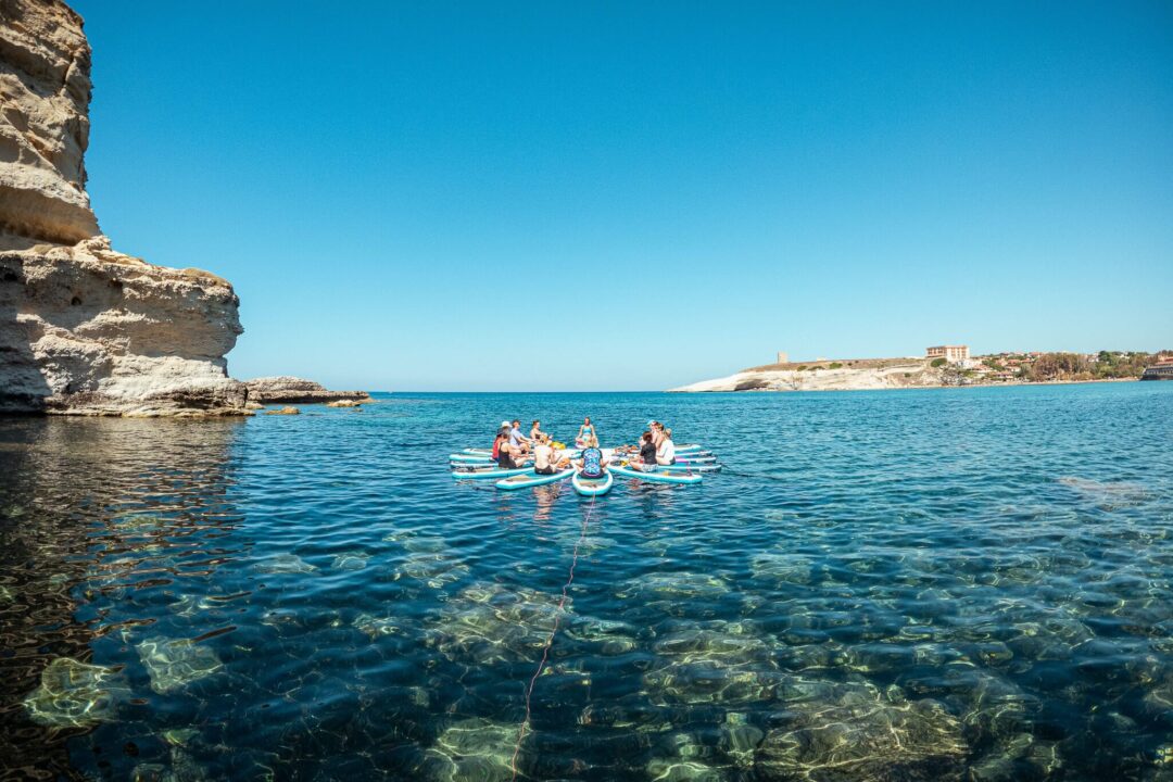 Women paddleboarding on clear sea near a rocky coast and distant beach under a blue sky