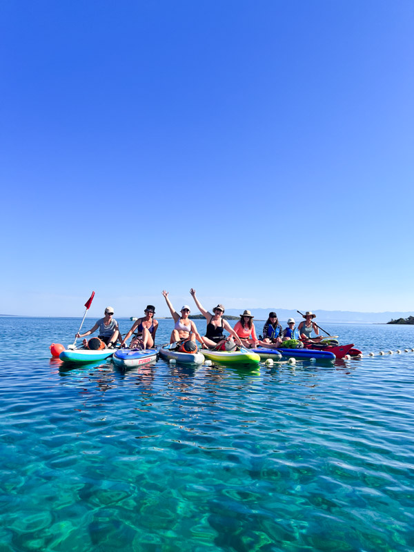 Women on paddleboards in clear blue sea, gathered for an island-hopping adventure by the coast