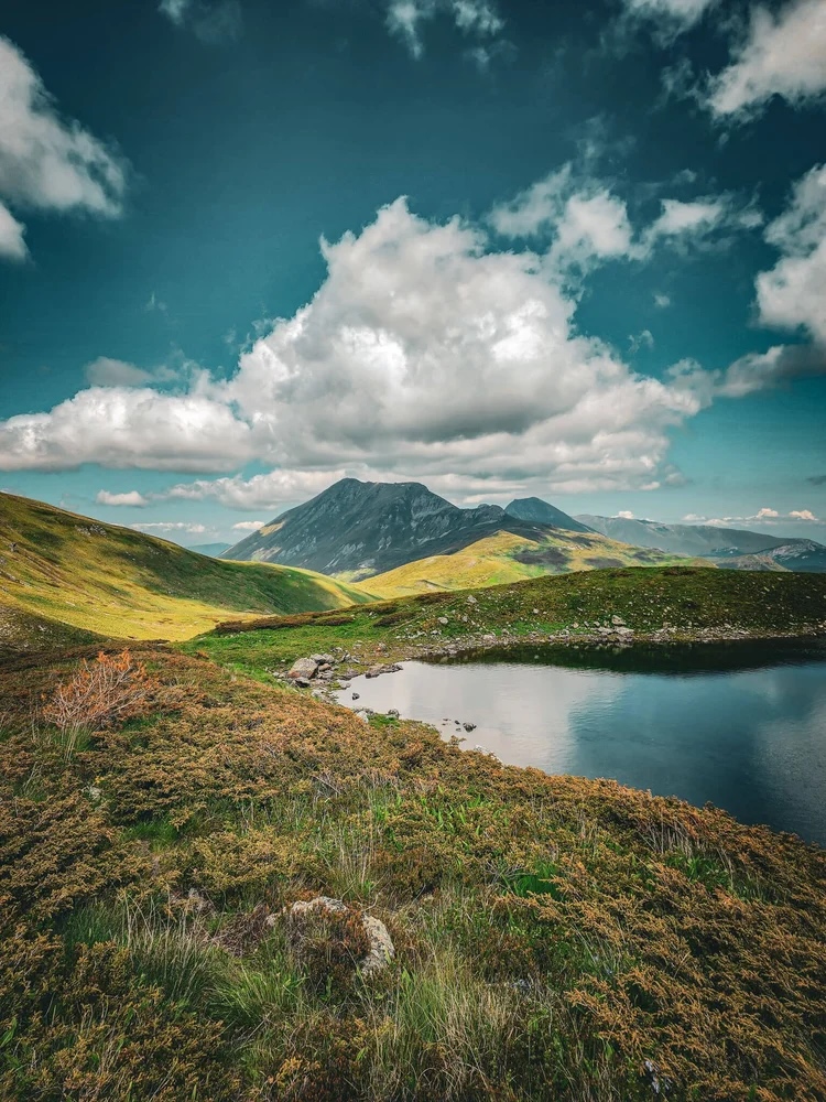 Mountain hiking landscape with a calm lake, grassy slopes and dramatic clouds under peaks