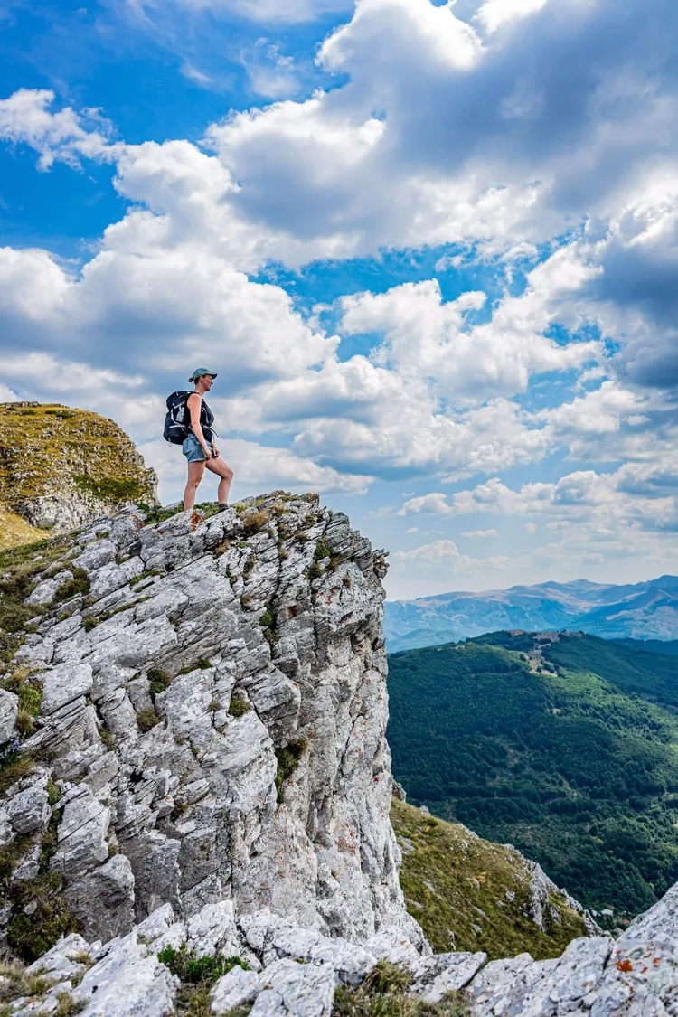 Woman hiking on a rocky ridge above forested valleys in North Macedonia under a cloudy blue sky
