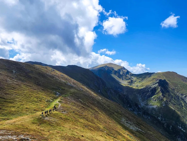Mountain hiking trail with hikers on a grassy ridge under bright blue skies and cumulus clouds