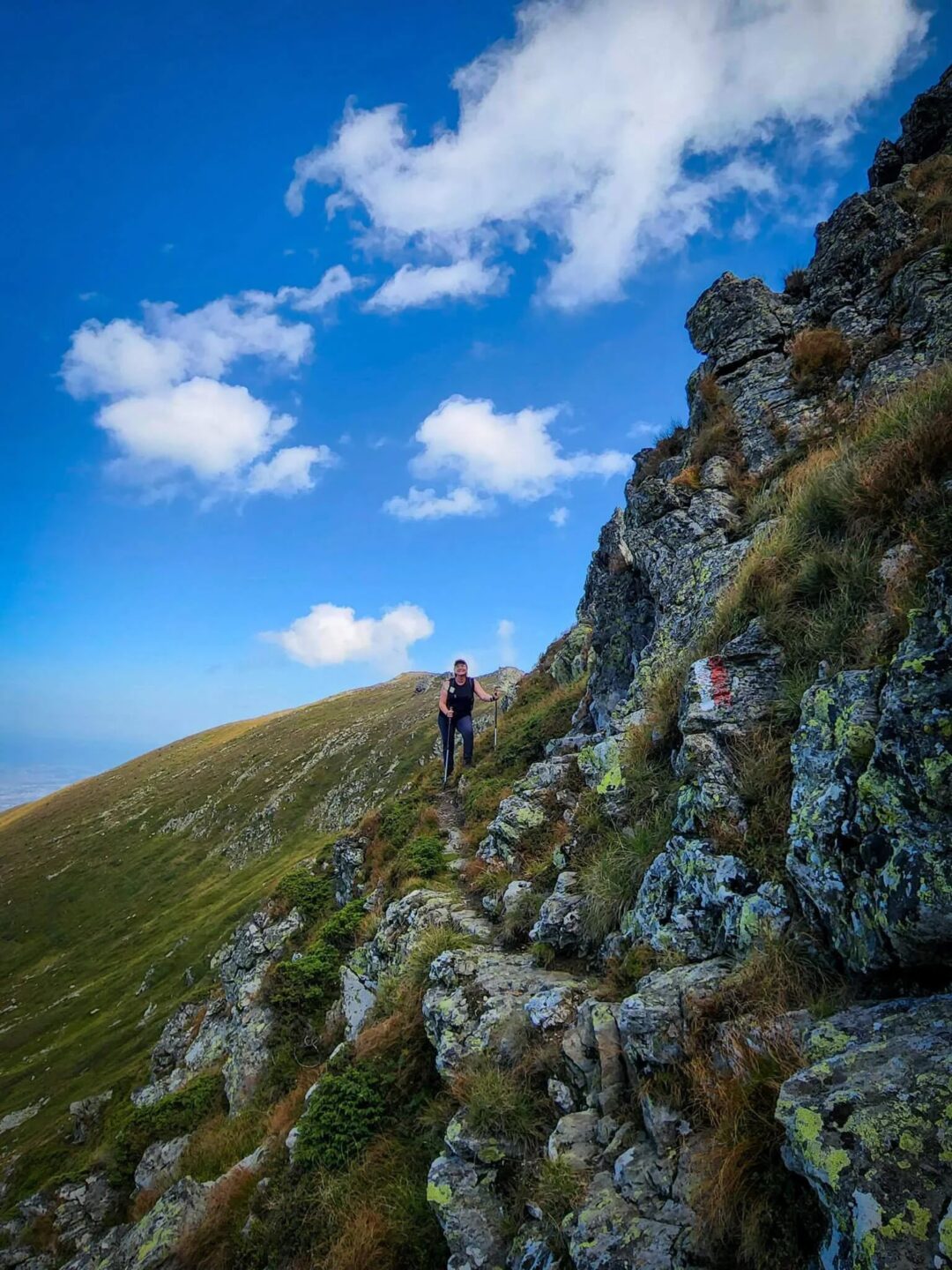 Woman hiking along a rocky mountain trail under blue skies and clouds on a scenic hillside.