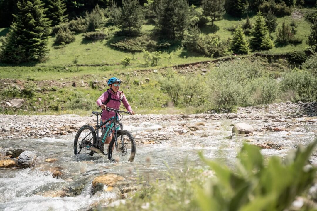 Woman e-biking through a rocky stream in a green mountain landscape