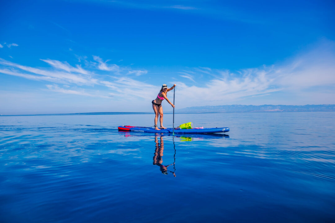 Woman paddling on a stand-up paddleboard on calm blue water with distant shoreline and mountains