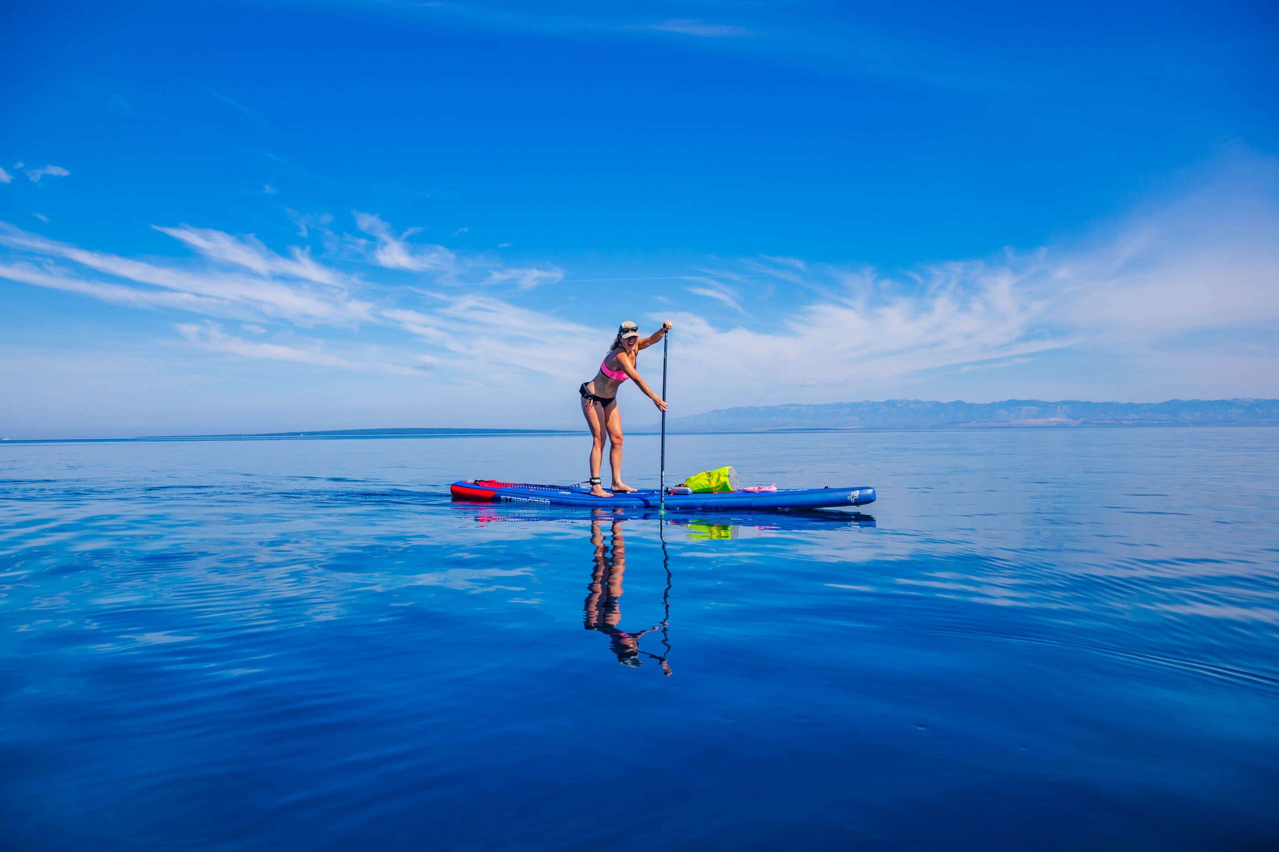 Woman paddling on a stand-up paddleboard on calm blue water with distant shoreline and mountains