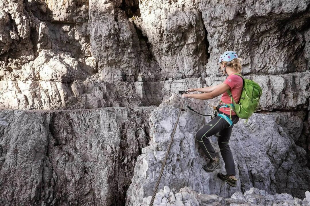 Woman hiking a via ferrata route on a rocky cliff face in the mountains with a safety rope and harness