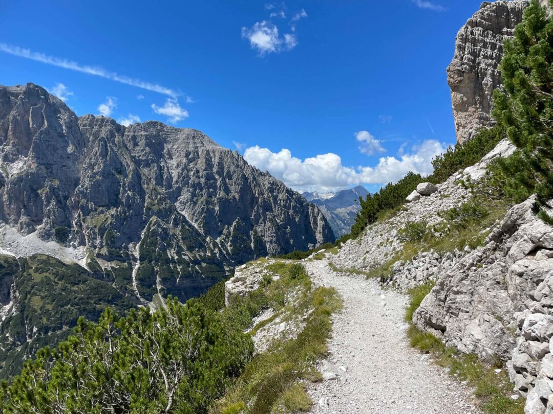 Mountain trail in the Dolomites with steep rock cliffs and forested slopes under a blue sky, multi-activity hiking route