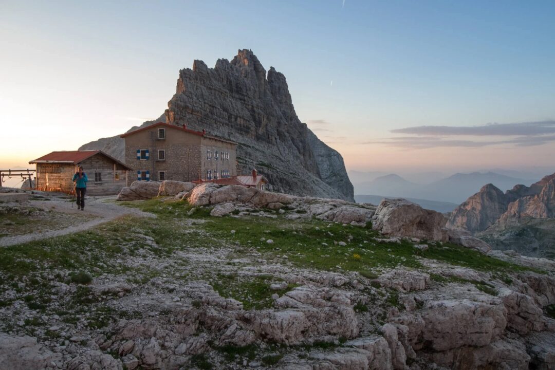 Woman walking past a mountain hut in the Dolomites under a rocky peak at dusk