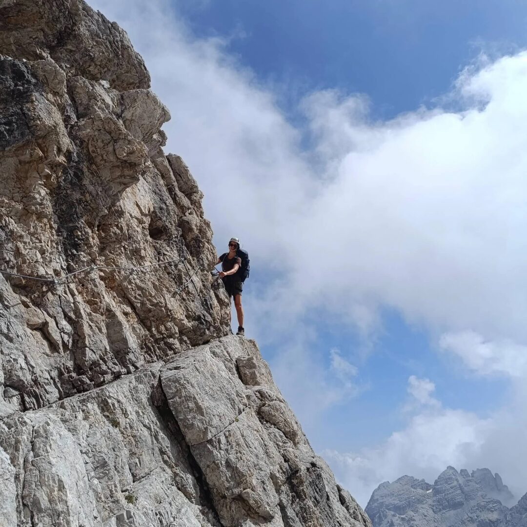 Woman standing on a rocky cliff edge in alpine mountainous terrain, surrounded by cloud and peaks