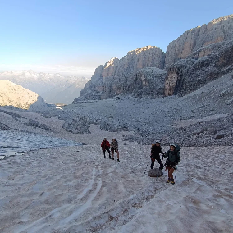 Hikers on a snowy mountain trail with backpacks in the Dolomites, passing high rock faces at dawn