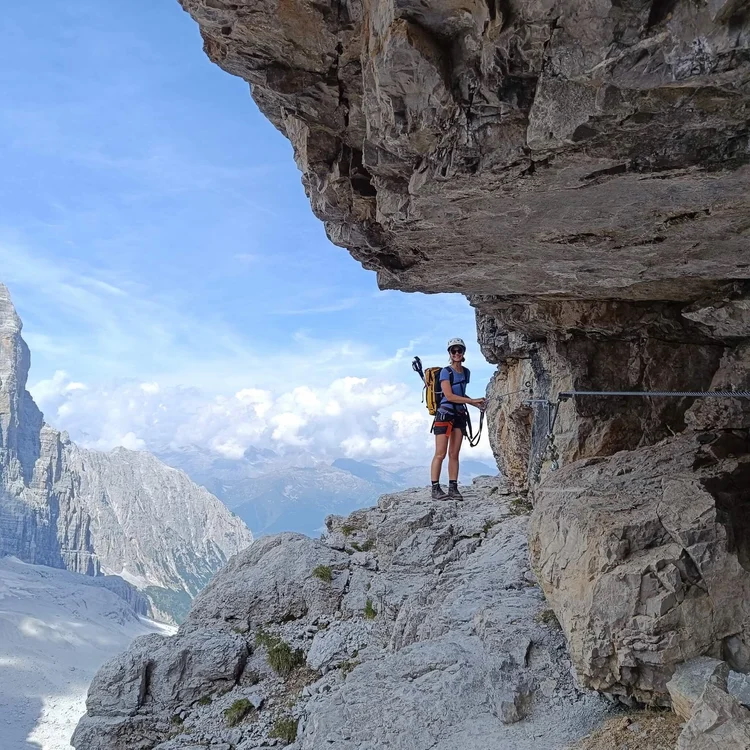 Woman hiking a via ferrata on a steep rock ledge in the mountains, wearing a safety harness and backpack