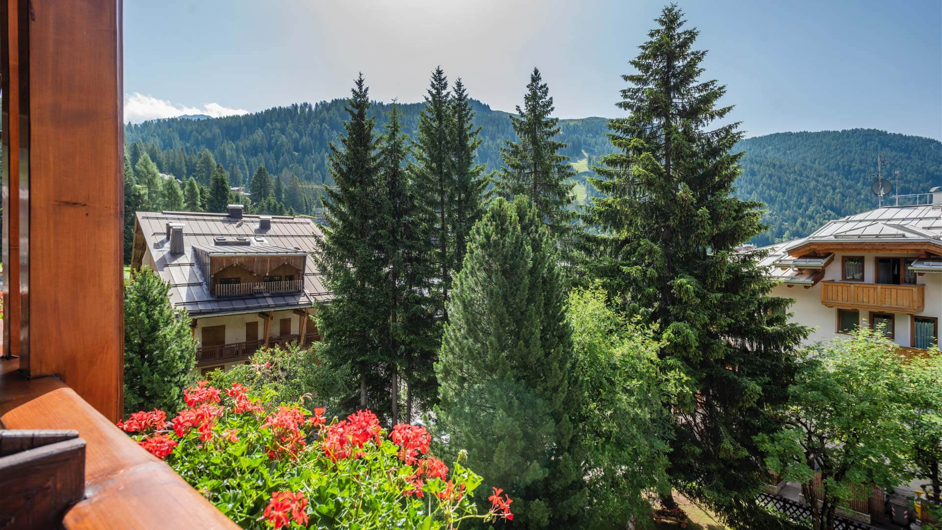 Dolomites mountain hut view from a balcony with pine trees, flower beds and timber chalets under blue sky
