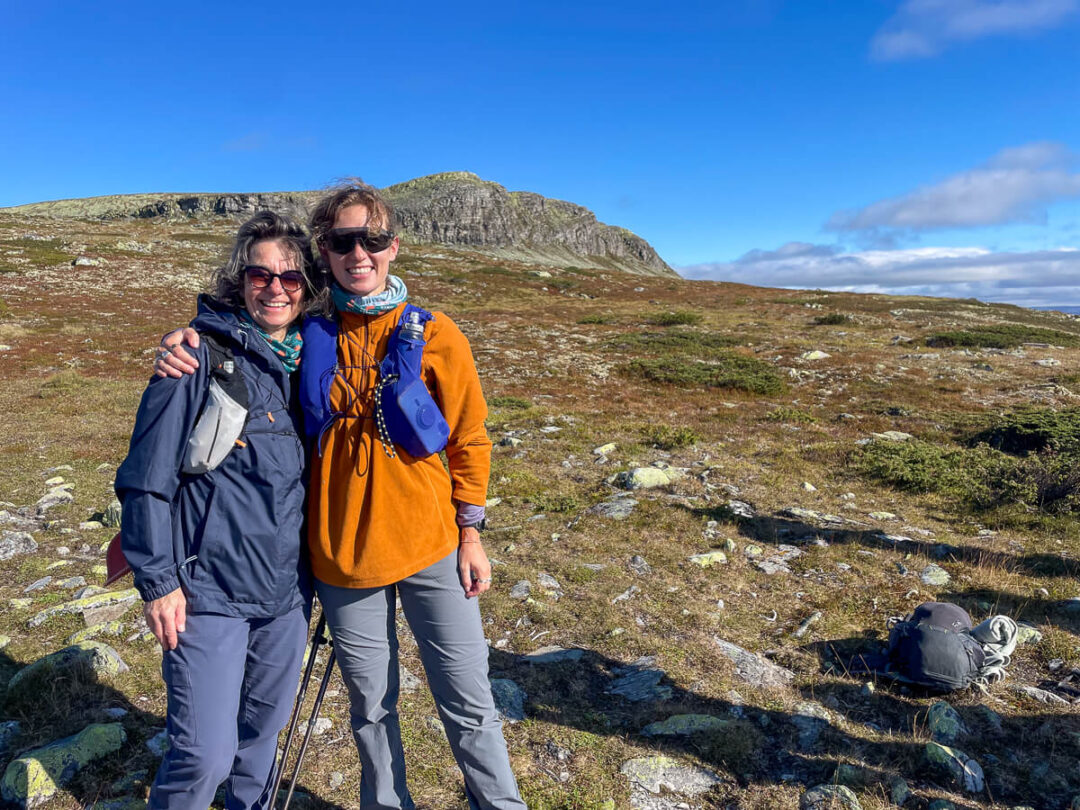 Two women hiking with backpacks on a rocky mountain trail in Norway under blue skies