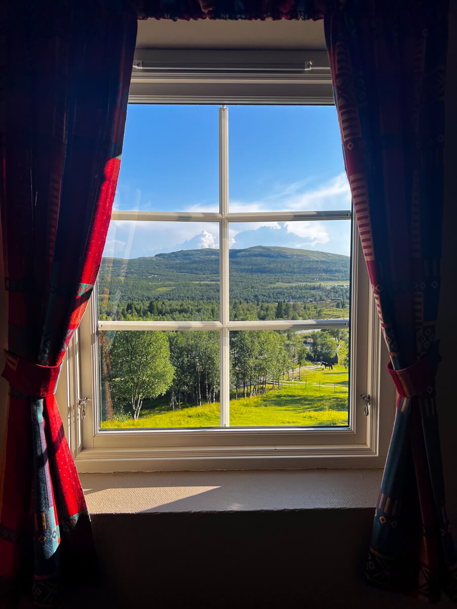 Scenic mountain valley view through a window with patterned curtains, framed greenery and hills in the distance