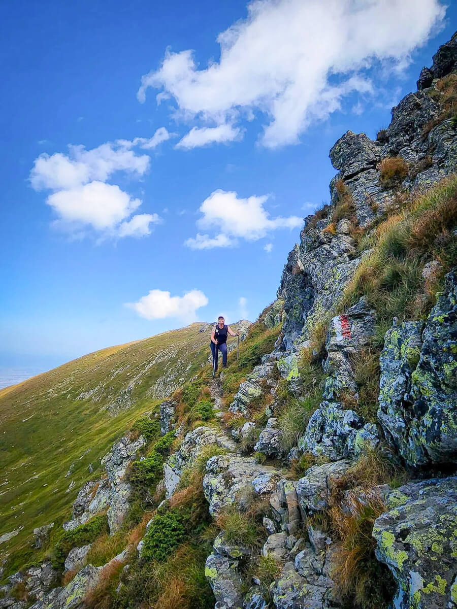 Woman hiking on a rocky mountain trail with grassy slopes under a blue sky and clouds