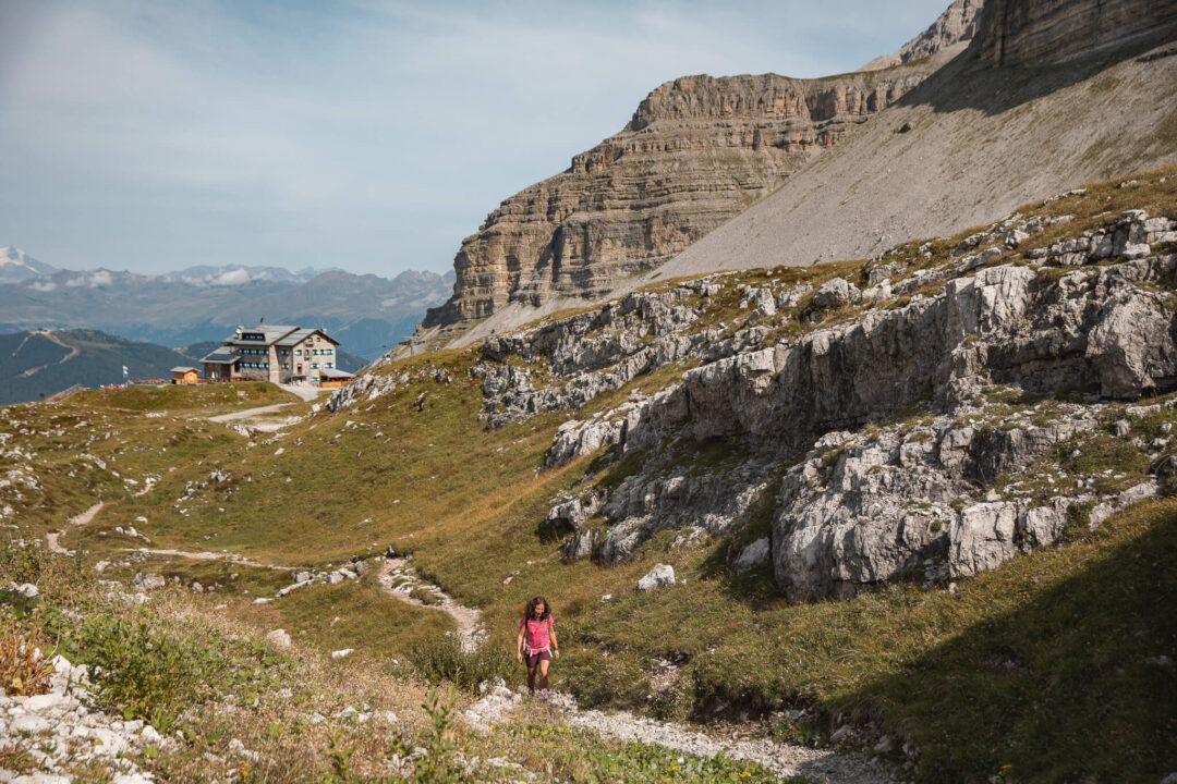 Woman hiking a mountain trail past an alpine lodge in rocky alpine countryside