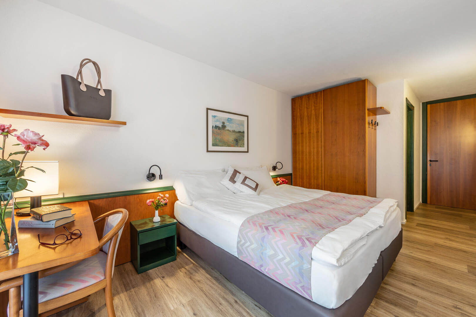 Bright hotel bedroom with single bed, desk and wardrobe, with framed wall art and warm lighting.