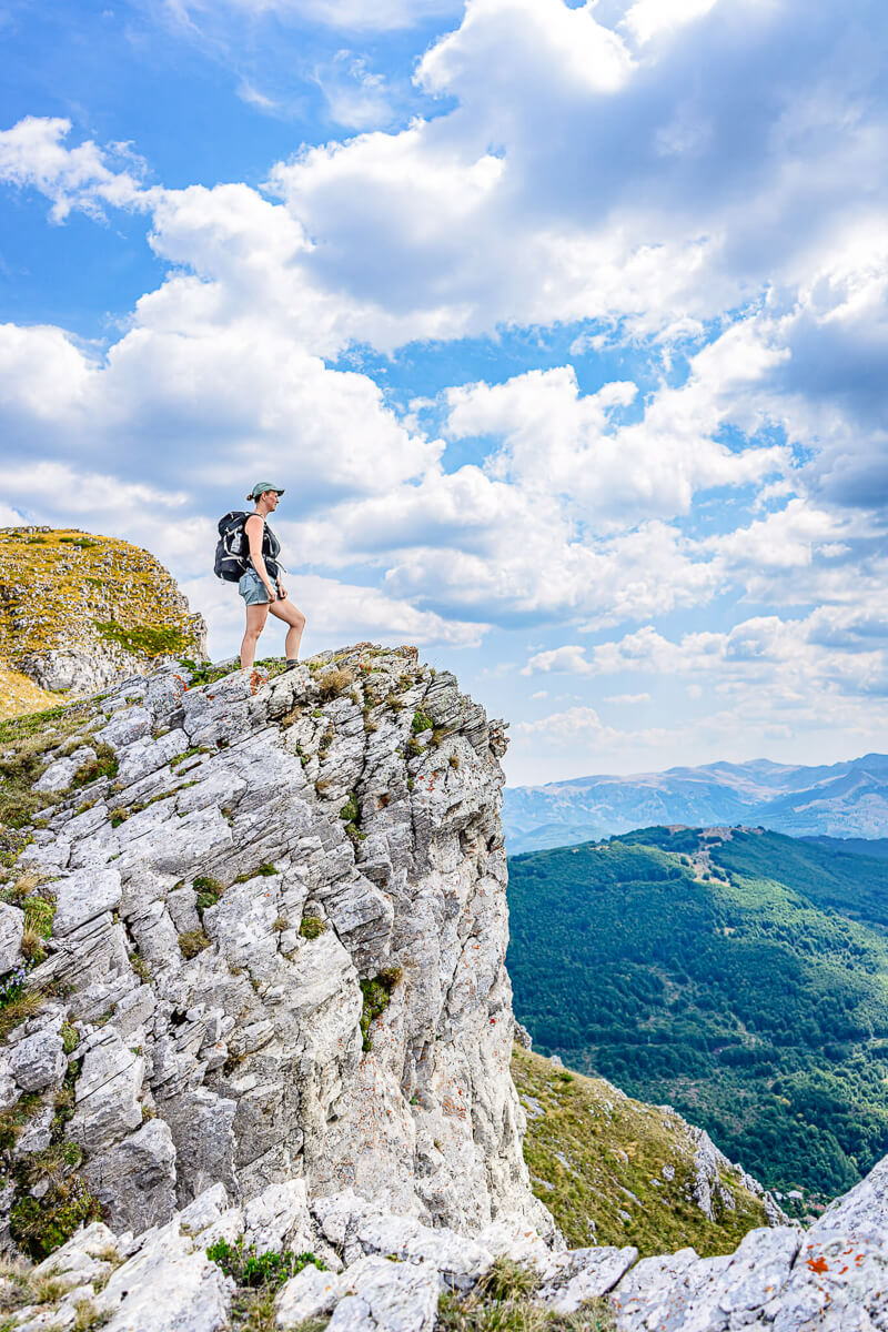Woman hiking on a rocky cliff with mountain valley views under a blue, cloudy sky