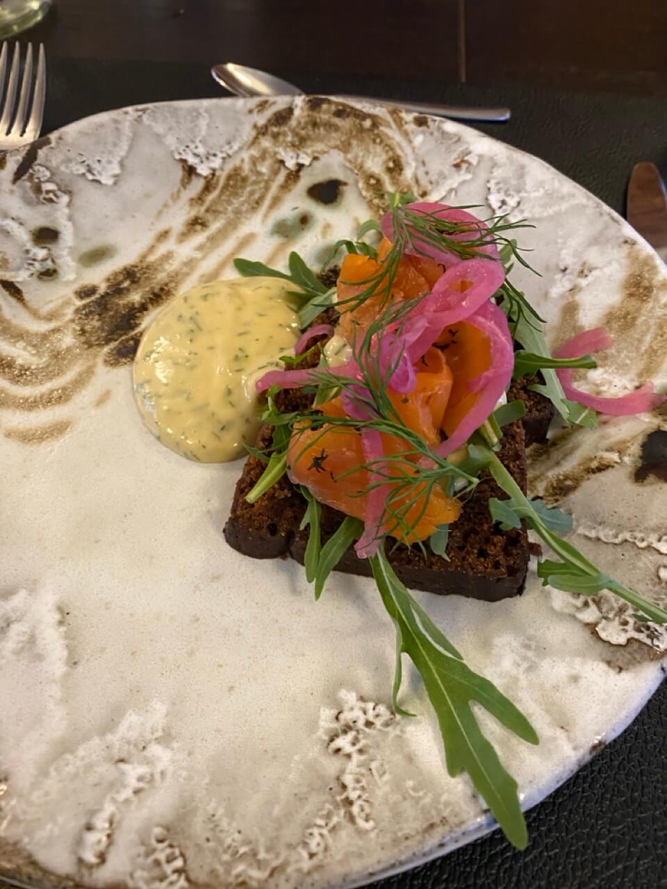 Open-faced starter on a plate with greens, edible flowers and pink pickled onions, served with sauce