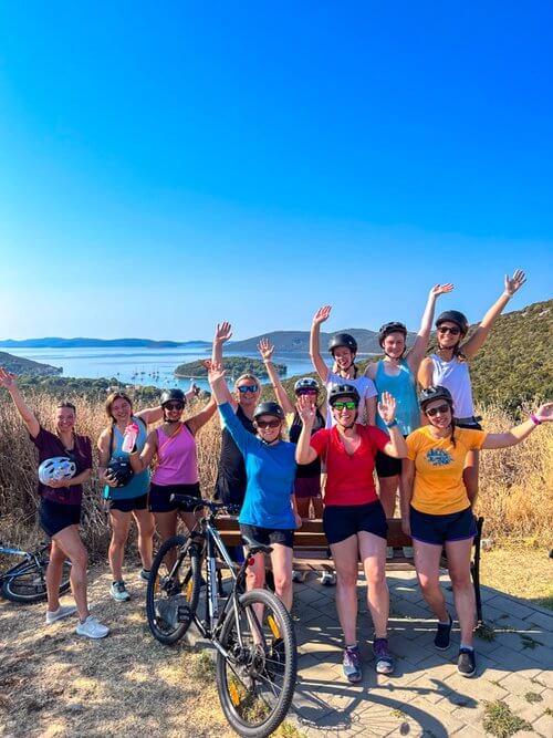 Women posing with bicycles on a coastal path overlooking the sea under a blue sky