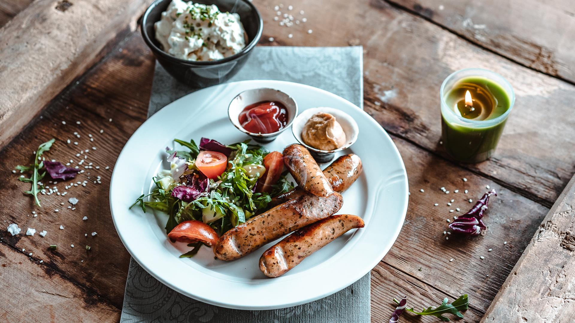 Sausages with salad and dipping sauces on a plate, with a lit candle on a wooden table outdoors