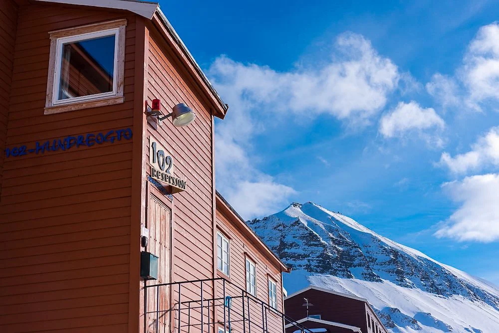 Red wooden cabin with balcony beside snowy mountains under a bright blue sky